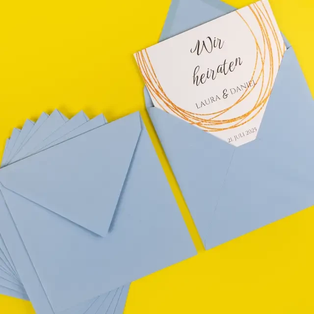 Eine quadratische Grußkarte mit der Aufschrift „Wir heiraten“ steckt im himmelblauen Umschlag. Daneben weitere Umschläge desselben Typs im 10er-Pack.