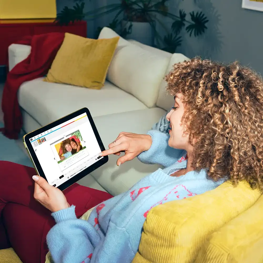 Einen Fotokalender bequem von zuhause aus selber machen Eine lockige Frau gestaltet einen Kalender im ORWO-Fotodesigner von zuhause aus, auf der Couch sitzend, mit einem Tablet und sieht dabei tiefenentspannt aus.