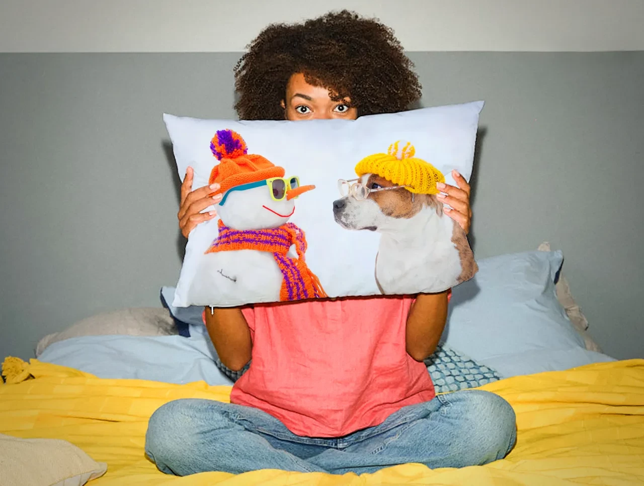 Auf einem gemütlichen Bett hält eine Frau mit dunkler Wuschelmähne ein großes Fotokissen spielerisch vor ihr Gesicht.