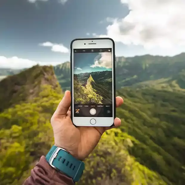 Fotografieren in den Bergen: Wandern und Erinnerungen festhalten Eine grün bewachsene Berglandschaft im Hintergrund, während ein Wanderer in Nahaufnahme seine Hand mit Smartphone hält und ein Foto macht. Der Moment wird eingefangen, um die atemberaubende Natur zu dokumentieren und die Erinnerung an den Ausflug festzuhalten.