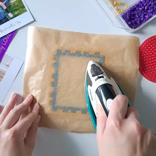 Bügelperlen-DIY: Bügeln des selbstgemachten Bilderrahmens Eine Person verwendet ein kleines Bügeleisen, um für das Bügelperlen-DIY durch Backpapier hindurch einen Bilderrahmen aus bunten Bügelperlen zu verschmelzen.