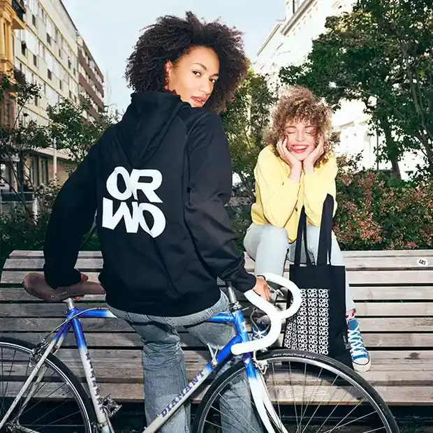 ORWO Merchandise in Aktion Zwei junge Frauen genießen die frische Luft. Eine sitzt auf einem Fahrrad und trägt einen stylischen ORWO-Hoodie, während die andere auf einer Bank sitzt, lacht und stolz eine ORWO-Stofftasche präsentiert – perfekte Begleiter für den Alltag.