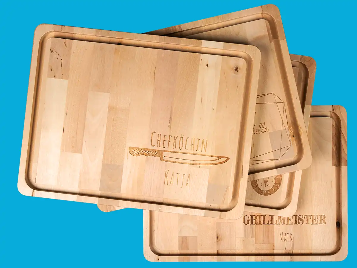 Mehrere gravierte Holzbretter für die Küche, übereinander gestapelt. Eine zeigt die Gravur „Chefkoch Katja“ mit einem Messer als grafisches Element, eine andere „Grillmeister Maik“. Diese gravierten Geschenke sind perfekte, persönliche Highlights für Hobbyköche und Grillfreunde.