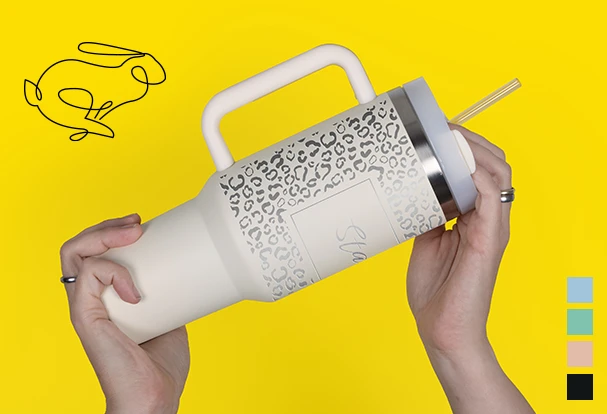Oster Geschenkideen: Ein großer, personalisierter Thermobecher im Stanley-Style wird von zwei Händen in die Höhe gehalten. Die Gravur auf dem Becher macht ihn zu einem einzigartigen Geschenk.