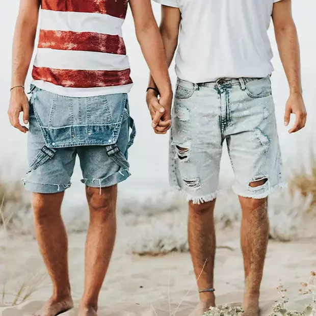 Tipps für schöne Strandfotos – Romantische Momentaufnahmen Ein Paar steht Hand in Hand am Strand, nur die Beine ab Hüfte abwärts sind zu sehen. Die sommerliche Kleidung und der Sand unter ihren Füßen schaffen eine entspannte, romantische Atmosphäre – eine wunderbare Inspiration für Strandfotos.