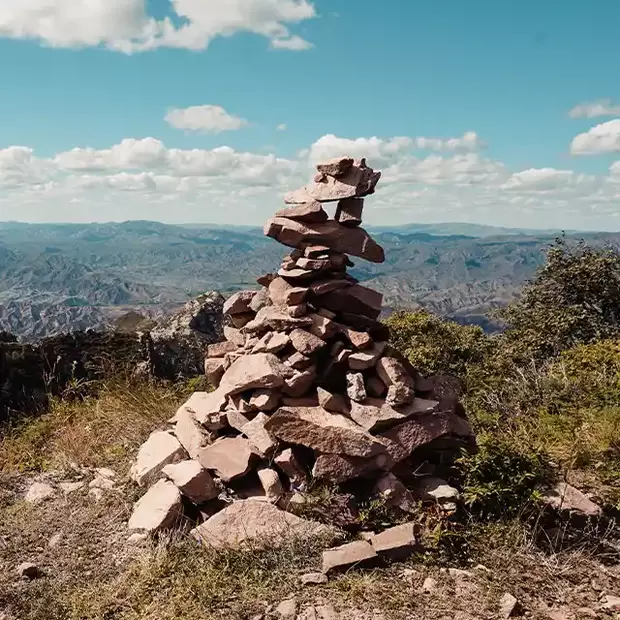 Fotografieren in den Bergen: Steintürmchen über dem Tal Ein beeindruckender Blick ins Tal, mit einer locker gestapelten Pyramide aus unterschiedlich großen Steinen am Rand eines Abgrunds. Der strahlend blaue Himmel mit weißen Wolken bildet einen faszinierenden Kontrast zu den natürlichen Elementen, die die Berglandschaft umrahmen.