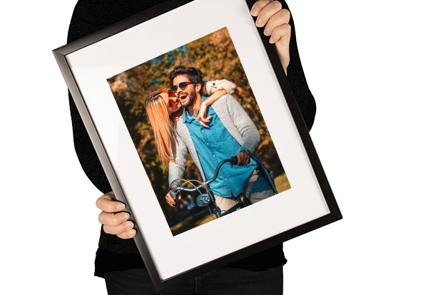 Person hält einen schwarzen Bilderrahmen mit einem Foto eines glücklichen Paares auf einem Fahrrad – personalisierbares Fotoprodukt von ORWO.