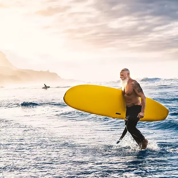Tipps für schöne Strandfotos – Magische Sonnenuntergangsmomente Ein Surfer mit auffälligem weißem Bart kommt aus dem Wasser, sein Surfboard in der Hand. Der Hintergrund zeigt einen atemberaubenden Sonnenuntergang, der die Szenerie in warmes Licht taucht – eine perfekte Aufnahme für beeindruckende Strandfotos.