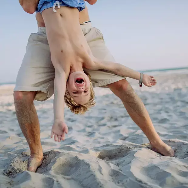 Tipps für schöne Strandfotos – Spaßvolle Momente am Meer Ein Junge wird kopfüber an den Beinen gehalten, während er lachend am Strand schwebt. Die Nahaufnahme fängt den Spaß und die Freude der Szene ein – eine lebendige und dynamische Momentaufnahme für Strandfotos voller Energie.