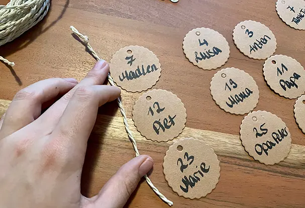 Die Pappschildchen liegen geordnet untereinander, bereit für die nächsten Schritte. Die Schnur wird gezeigt, wie sie an den Schildchen befestigt wird, um später die Namen der Geburtstagskinder anzuhängen. Dieser Schritt ist ein wichtiger Teil des DIY-Prozesses für deinen individuellen Geburtstagskalender aus Holz.