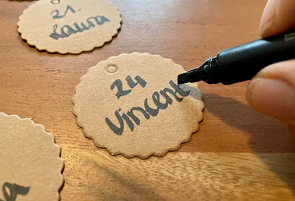 Nahaufnahme eines Pappschildchens, das mit einem Namen für den DIY Geburtstagskalender aus Holz beschriftet wird. Die kleinen Schilder werden genutzt, um die Namen der Geburtstagskinder zu zeigen und so den Kalender persönlich und einzigartig zu gestalten.