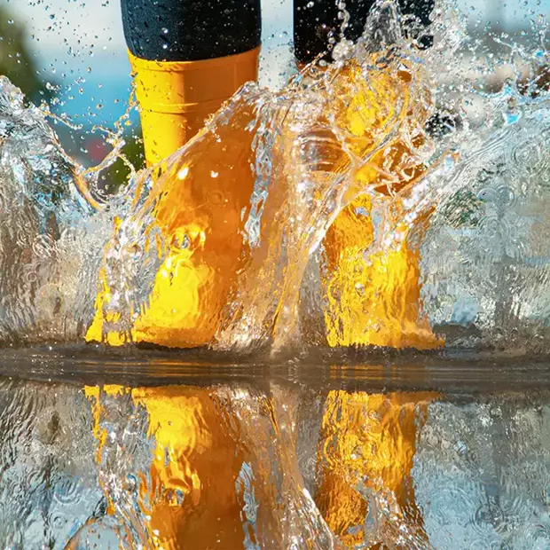 Eine Person mit gelben Gummistiefeln springt in eine Pfütze. Wasser spritzt nach oben. Auch bei Regenwetter kannst du schöne Frühlingsfotos machen.