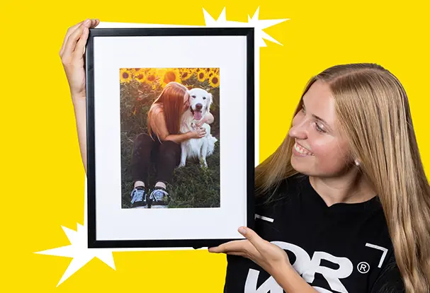Junge Frau hält lächelnd ein gerahmtes Foto von sich und ihrem Hund vor einem Sonnenblumenfeld vor einem leuchtend gelben Hintergrund. Bestell dein Lieblingsbild im Rahmen bei ORWO.