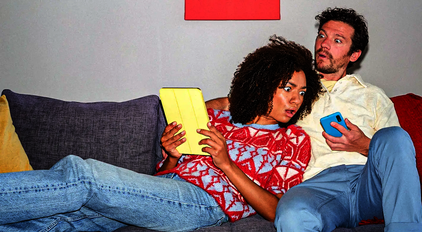 Valentinstag Fotogeschenke mit humorvollem Paarmotiv: Frau und Mann sitzen auf dem Sofa und schauen überrascht auf Tablet und Smartphone, modernes Alltagsmotiv für persönliche Fotogeschenke.