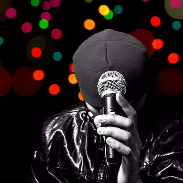 Minimalistisches Bild, aber durch einen Bokeh-Effekt professionell in Szene gesetzt. Ein schwarz gekleideter Sänger, in seiner Musik versunken, wird durch einen dunklen Hintergrund mit einem bunten Lichter Bokeh-Effekt professionell in Szene gesetzt.
