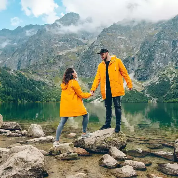 Bergfotos: Freunde am Bergsee Ein Paar in leuchtend gelben Mänteln steht vor einem kristallklaren Bergsee. Der Mann steht auf einem großen Findling und reicht der Frau die Hand, während die majestätische Berglandschaft im Hintergrund das romantische Szenario perfekt ergänzt.