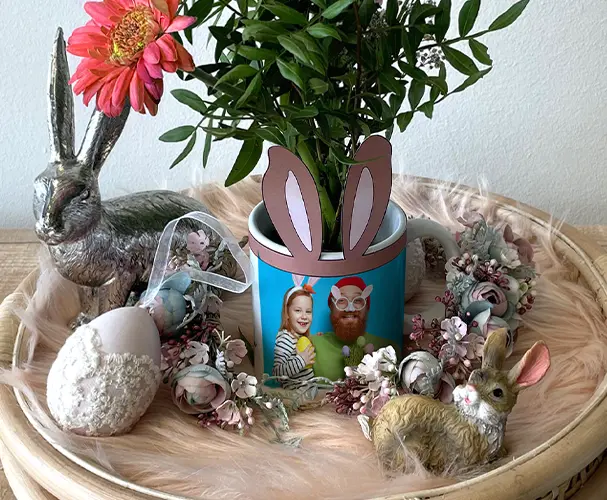 Eine kreativ gestaltete Fototasse dient als Blumentopf, dekoriert mit Osteraccessoires und Hasenohren aus Papier. Perfekt als Mitbringsel zu Ostern oder für personalisierte Ostergeschenke.