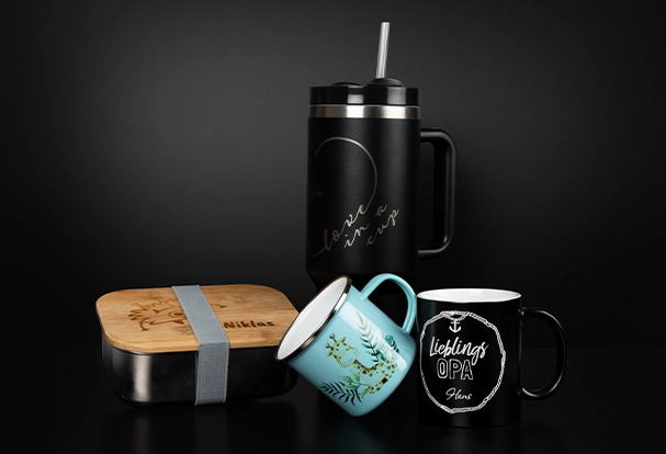 Top Angebote zum Black Friday. Vor schwarzem Hintergrund präsentieren sich einzigartige Fotogeschenke: ein gravierter XXL-Thermobecher, eine personalisierte Tasse, eine bedruckte Emailletasse mit kindlichem Motiv und eine Lunchbox mit individuellem Holzdeckel.