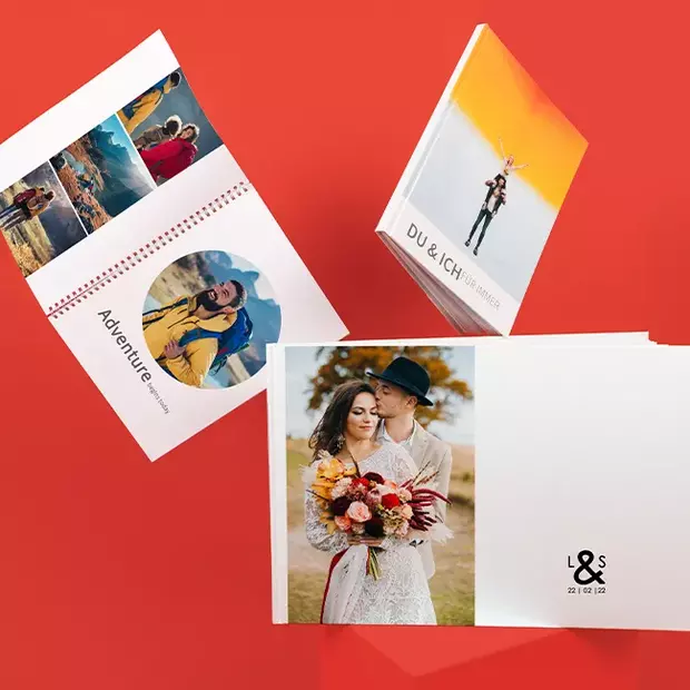 Drei Fotobücher in kreativer Anordnung: Hochzeitsbuch, Wanderbuch mit Ringbindung und Paarbuch „Du & ich für immer“, roter Hintergrund.