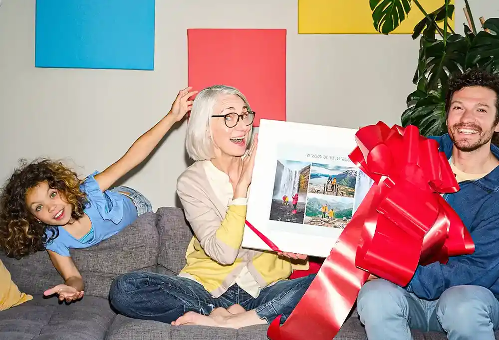 Oma, Papa und Enkelin sitzen auf der Couch. Oma bekommt ein Fotoprodukt mit großer, roter Schleife von Ihrem Sohn.
