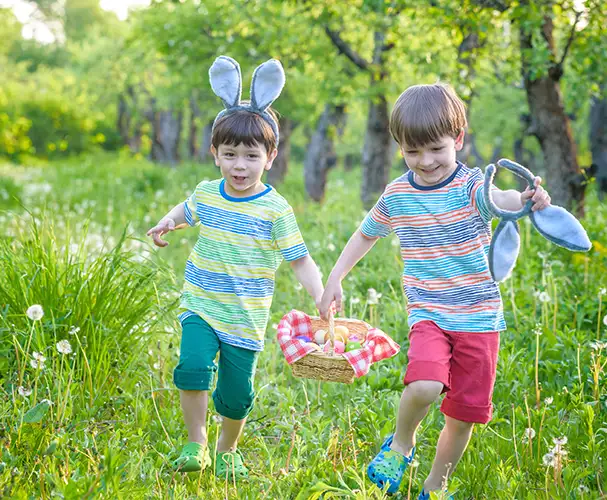 Zwei Kinder, die einen Korb voller bunter Ostereier über eine Wiese tragen. Hol dir Inspiration für Frühlingsfotos zu Ostern.
