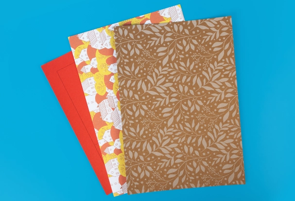 Drei verschiedene Bastelbögen für kreative Geschenkverpackungen. Ein roter Bogen, ein Muster mit kleinen Häusern in Rot, Gelb, Weiß und Schwarz sowie ein weißes Blumenmuster auf Kraftpapier.