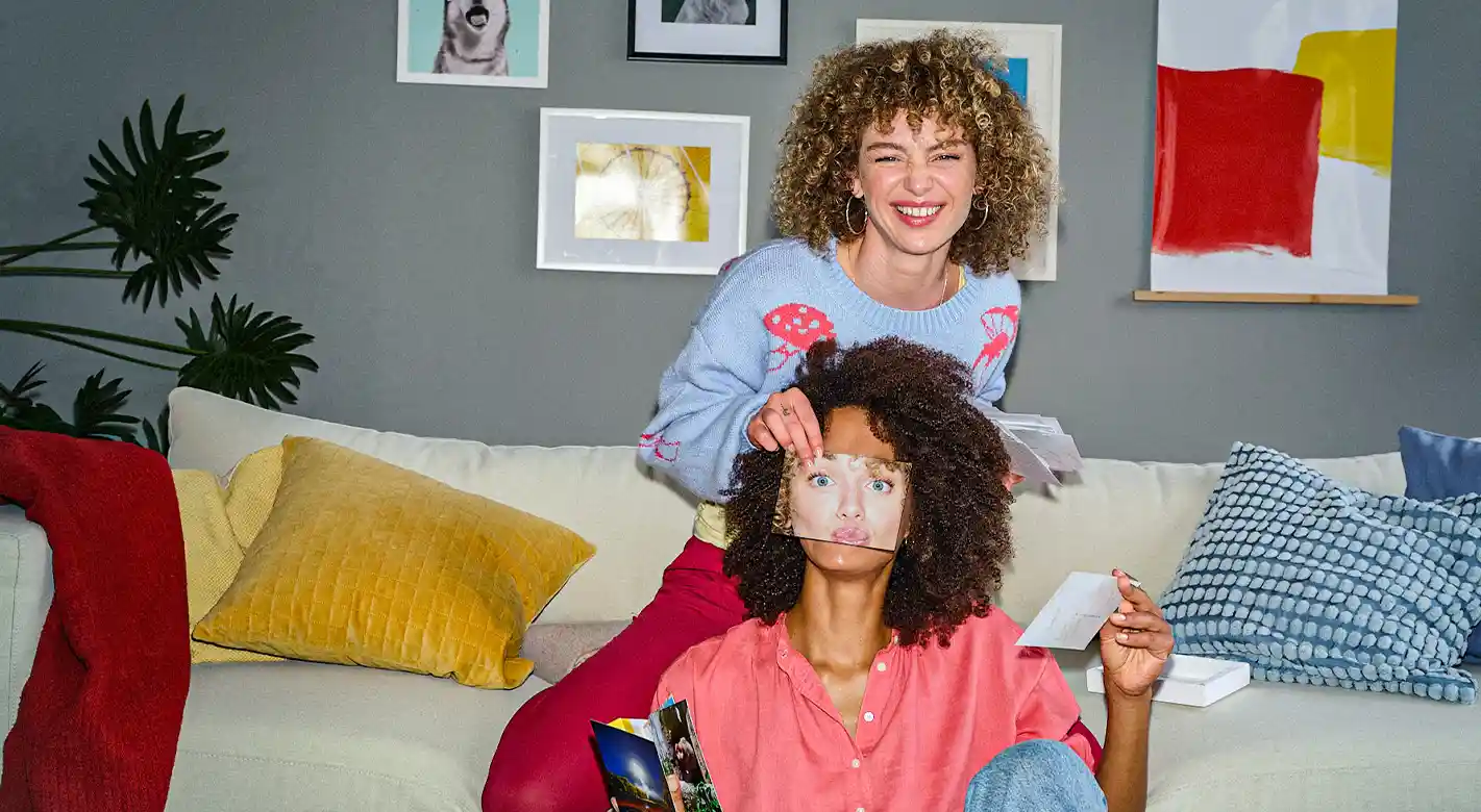Zwei junge Frauen haben Spaß mit Fotoabzügen. Eine hält ihrer Freundin ein Foto mit einem Gesicht vors Gesicht – als witzige Verkleidung. Das Wohnzimmer ist gemütlich eingerichtet mit heller Couch und farbenfrohen Wandbildern, die eine kreative Atmosphäre schaffen.