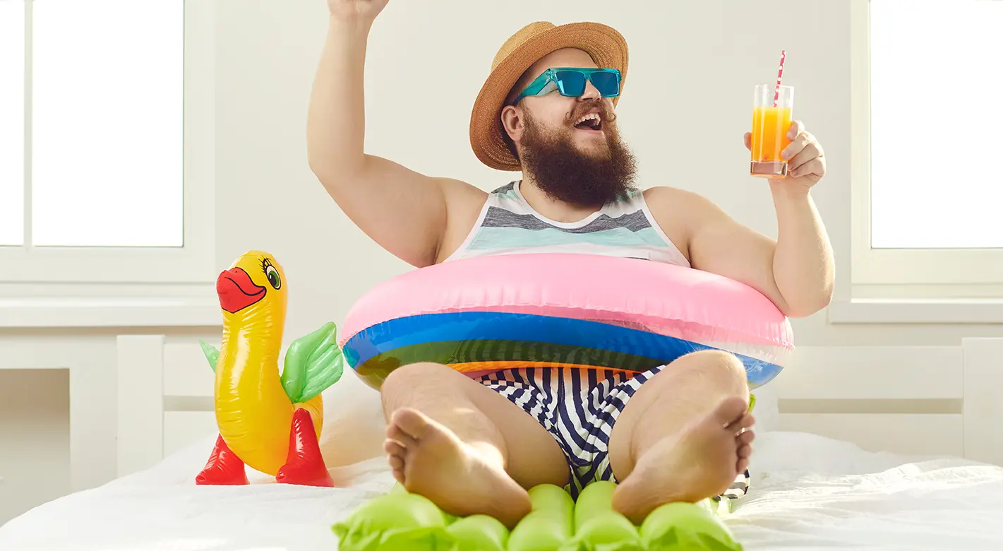 Ein Mann in Shorts, Hemd und Sonnenhut sitzt schwungvoll auf einer Luftmatratze, die auf einem Bett liegt. Mit einem Drink in der Hand und einer gelben Schwimm-Ente neben sich, vermittelt die Szene sommerliches Urlaubsfeeling – perfekt für kreative Strandfotos.