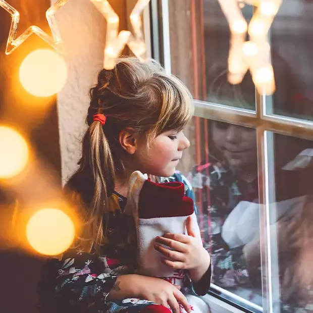 Geschichte zum Adventskalender Ein kleines Mädchen sitzt mit einer Wärmflasche am Fenster und blickt nach draußen, umgeben von einer gemütlichen, weihnachtlichen Atmosphäre. Der Moment spiegelt die warme Vorfreude auf den kommenden Advent wieder, die Geschichte eines Weihnachtskalenders in einem Bild eingefangen.