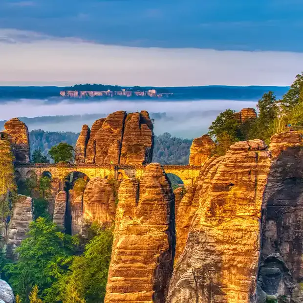 Licht und Schatten auf einem Foto mit Langzeitbelichtung: Während die Morgensonne Berge und Brücke beleuchtet umhüllt ein mystischer Nebel den Wald im Hintergrund.