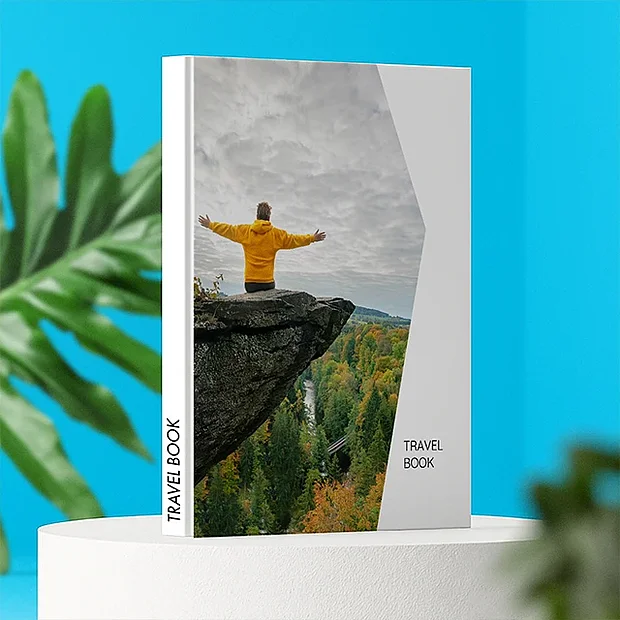 Hochformatiges Fotobuch mit Reisebild einer Person auf einem Felsen über einer Landschaft, präsentiert auf weißem Podest vor blauem Hintergrund