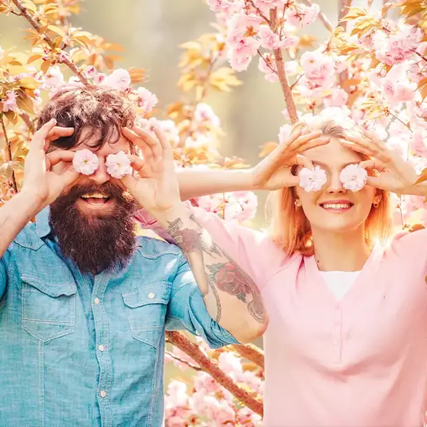 Ein Mann und eine Frau stehen vor einem rosa blühenden Baum und halten sich jeweils zwei Blüten vor die Augen. Blüten sind tolle Requisiten für Frühlingsfotos.