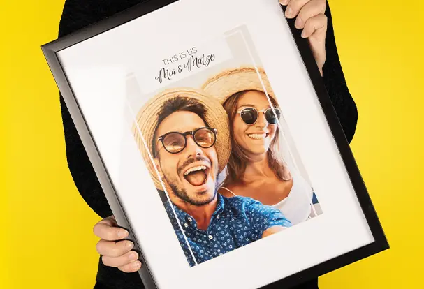 Ein Poster mit einem Bild von einem Paar in Strohhüten, das den Schriftzug "Mrs. & Mrs." trägt, im Bilderrahmen. Die Präsentation erfolgt durch eine Frau, die das Erinnerungsstück zeigt. Um das Ambiente zu ergänzen, könnten auch Sternzeichen-Wandbilder eine wunderbare Ergänzung für deine Wanddekoration sein.
