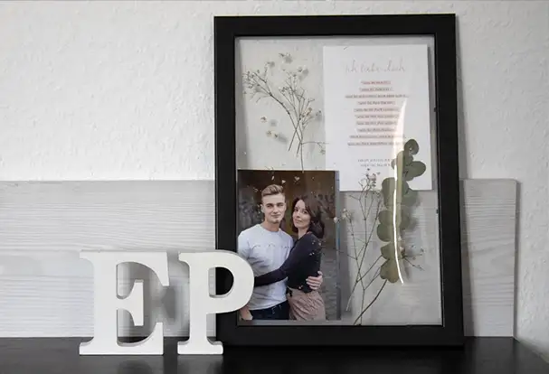 Ein DIY Bilderrahmen mit zwei Glasscheiben, ohne traditionelle Rückwand. Zwischen diesen Glasscheiben sind ein Foto, ein geschriebener Text und Trockenblumen zu sehen.