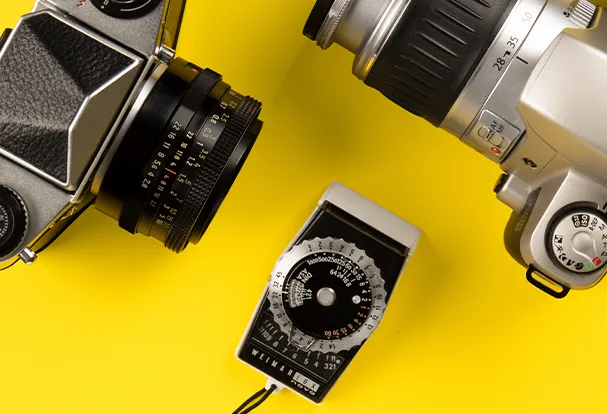 Es werden ein klassischer Belichtungsmesser und zwei analoge Spiegelreflexkameras aus vergangenen Zeiten gezeigt.