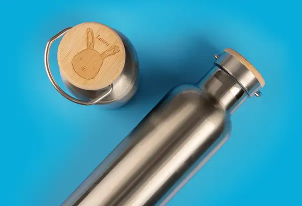 Eine silberne, metallische Thermosflasche mit einem gravierten Holzdeckel, personalisiert mit einem Namen und einem süßen Hasenmotiv. Perfekt als Gravur Geschenk für Kinder, das sowohl praktisch als auch einzigartig ist.