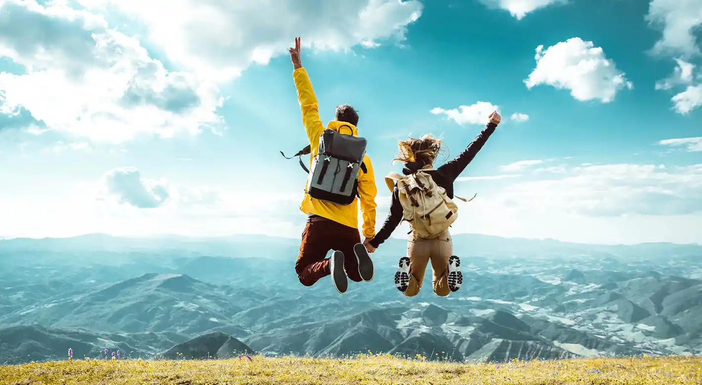 Ein Paar springt jubelnd in die Luft auf einer Wandertour und genießt die atemberaubende Aussicht auf das Tal und die umliegenden Berge. Das Bild fängt den Moment der Freude und den unvergesslichen Blick auf die majestätische Berglandschaft ein.