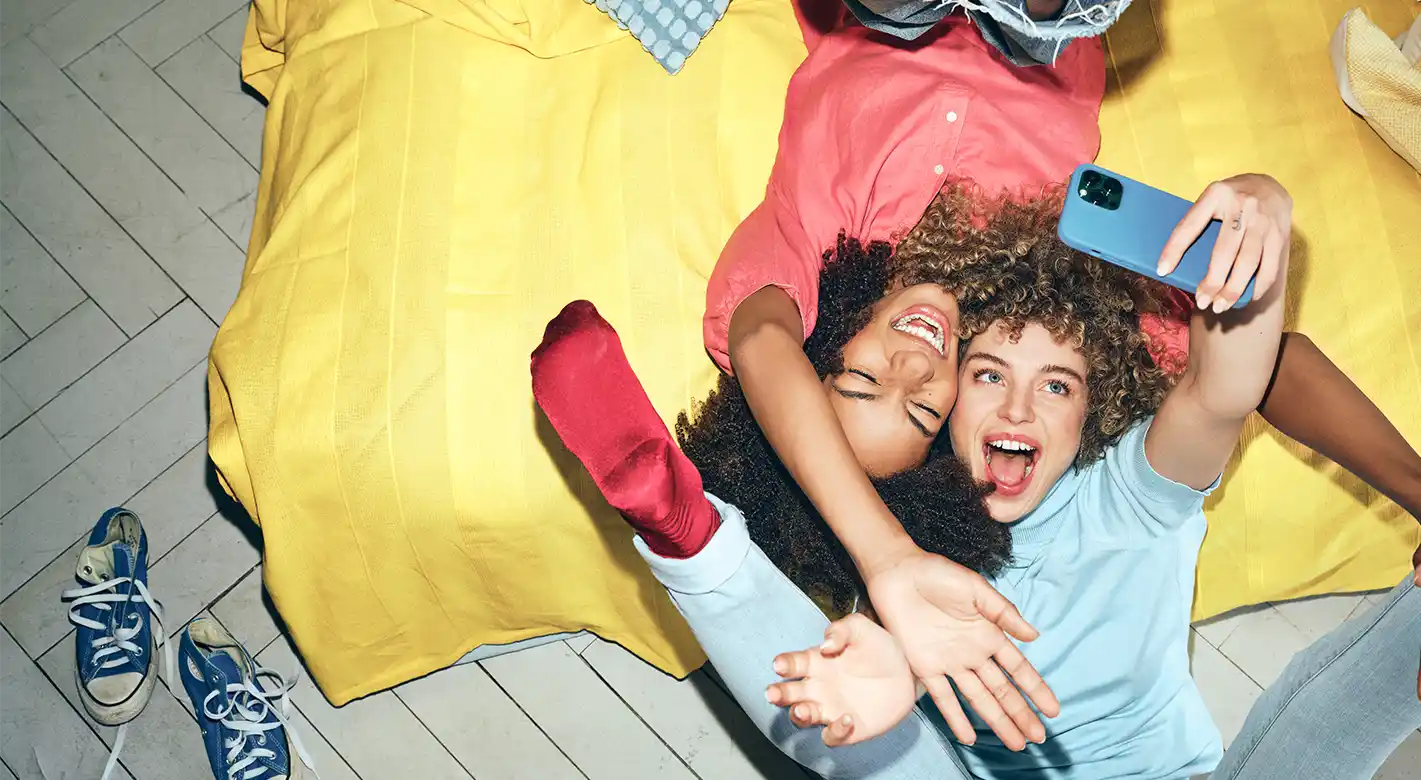 Zwei junge Frauen machen mit ihrer Foto-App lustige Selfies, während sie auf einer Couch oder einem Bett liegen und ihre Füße in die Luft strecken.