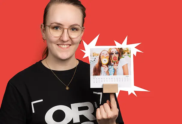 Junge Frau in ORWO Shirt hält lächelnd einen Tischkalender mit Foto hoch. Bestelle deinen eigenen Tischkalenderbei ORWO.