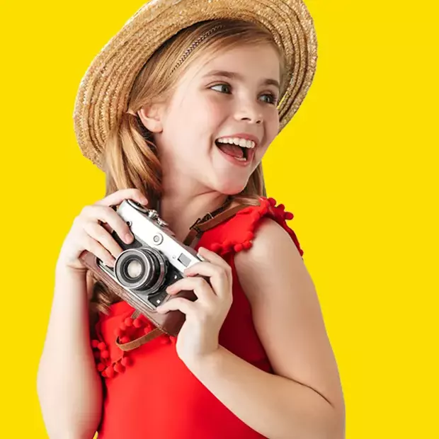 Mit Spaß fotografieren und analoge Filme entwickeln lassen – unvergessliche Momente auf Film Ein lachendes, blondes Mädchen im roten Sommerkleid mit Strohhut hält eine analoge Kamera freudig in den Händen.