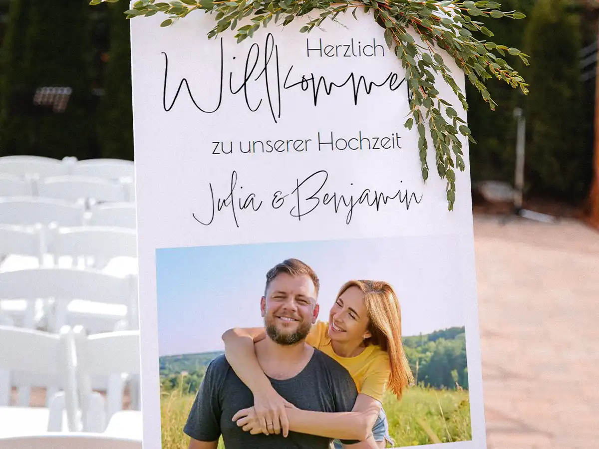 Eine große, weiße Hartschaumplatte, bedruckt mit einem Foto des Brautpaares und einem herzlichen Willkommensgruß für die Gäste. Das Design wird durch grüne Zweige geschmückt und setzt einen einladenden Akzent für den besonderen Tag.