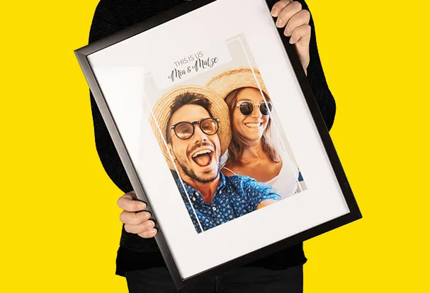 Eine Frau hält stolz einen schwarzen Bilderrahmen mit weißem Passepartout. Das Fotoposter darin zeigt ein glückliches Paar in Urlaubsstimmung. Perfekt für die stilvolle Dekoration mit persönlichen Erinnerungen.