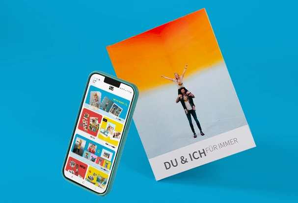 Smartphone mit Fotobuch-App neben personalisiertem Fotobuch mit Paarfoto und modernem Design auf blauem Hintergrund