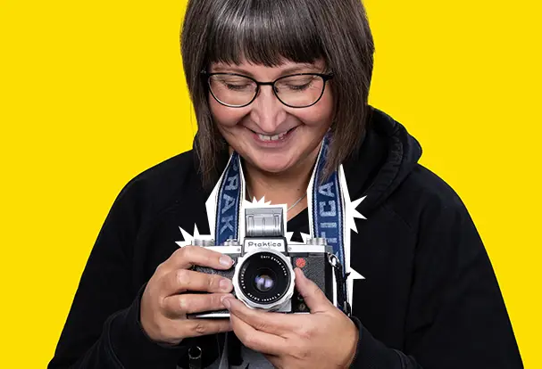 Analoge Fotografie mit Leidenschaft bei ORWO Lächelnde Frau mit Brille hält eine alte Praktica-Kamera vor gelbem Hintergrund. Entdecke die Faszination Analogfotografie mit ORWO.