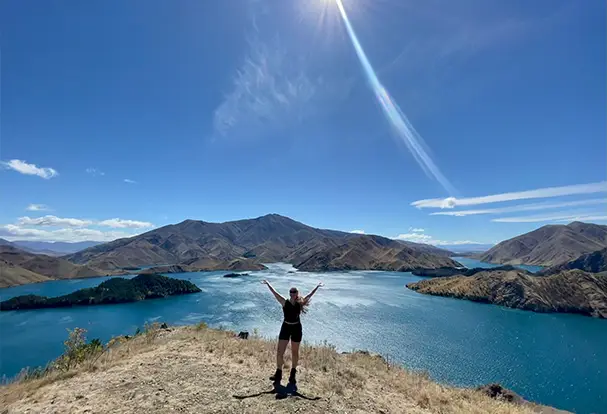 Freiheit und Abenteuer – Keesha gibt Urlaubsfototipps Junge Frau steht mit ausgebreiteten Armen auf einer Klippe mit Blick auf einen tiefblauen See und Berglandschaft unter strahlender Sonne. Lass deine Urlaubsfotos bei ORWO drucken!