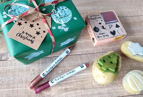 Individuelle Geschenkverpackung – vielleicht für ein Schneidebrett mit Lieblingsrezept Ein weihnachtlich verpacktes Geschenk in grünem Papier mit weißem Kartoffeldruck-Muster. Daneben eine geschnitzte Kartoffel mit Weihnachtsbaummotiv als Stempel.