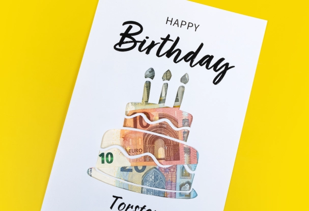 So kannst du stilvoll Geldgeschenke und Schokolade verpacken. Ein personalisiertes Poster zeigt eine Geburtstagstorte und die passenden Grüße. Es ist in einem weißen Rahmen eingefasst. Die Torte wurde ausgeschnitten und einige Geldscheine dahinter gelegt. Jetzt noch eine Tafel Schokolade verpacken und fertig ist dein Unikat