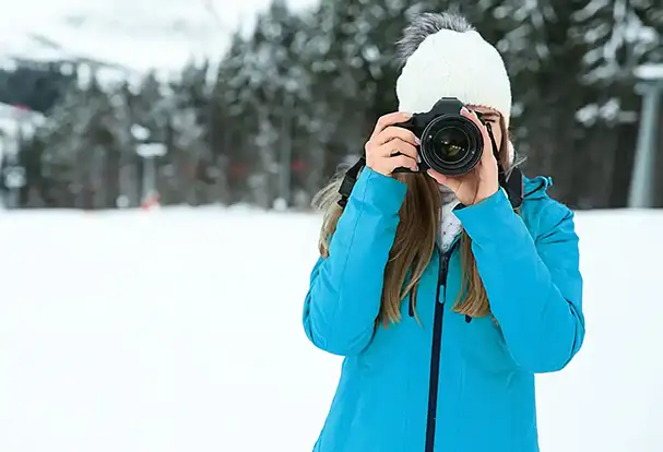 Fotografie-Tipps für beeindruckende Winterfotos Eine Frau in einer blauen Jacke fotografiert mit ihrer Kamera im Schnee. Entdecke jetzt unsere Foto Tricks für Winterfotos.