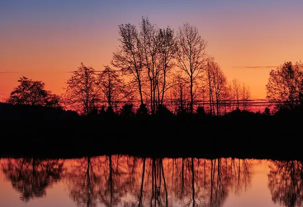 Magische Himmelsfärbungen spiegeln sich im Wasser Foto am See zur magischen Stunde. Die Bäume und Büsche am Uferrand erscheinen als schwarze Silhouette vor einem traumhaften orange- bis lilafarbenen Hintergrund welcher sich aus Himmel und Wasser zusammensetzt.