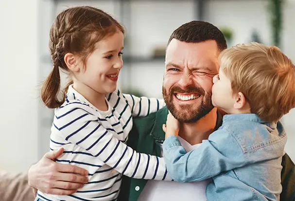 Fotografie Tipps – für emotionale Vatertagsfotos Strahlender Vater wird von seinen beiden Kindern umarmt und geküsst – Fotografie Tipps zum Vatertag.